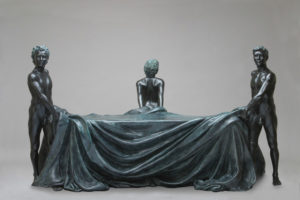 Entitled The Bench, this is a photograph depicting three one-quarter life-size bronze standing figures; two male and one female. They are holding a cloth draped over a bench, the male figures holding each end of the bench and the female stands at the back in the middle. Created by sculptor Blake Ward